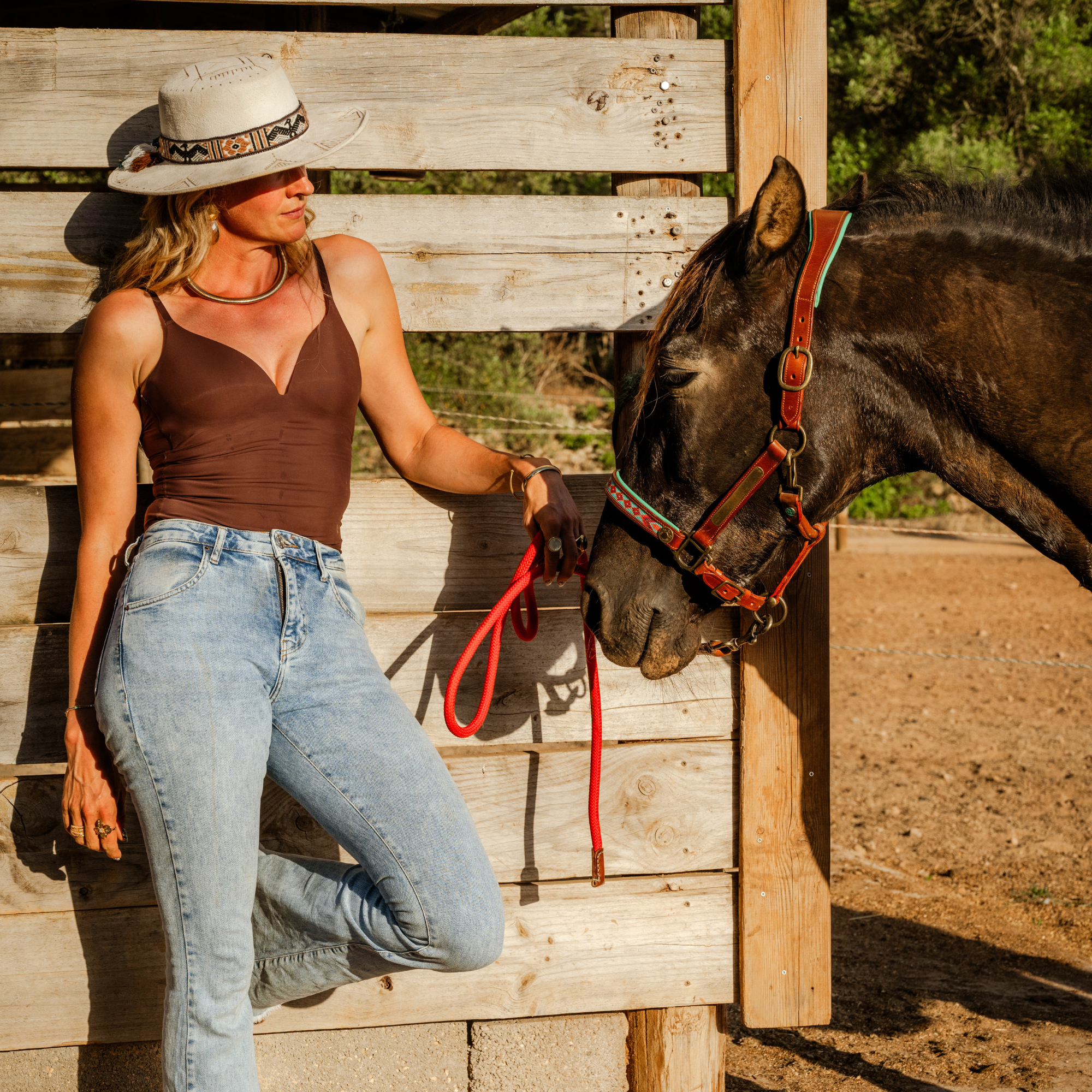 Woman in boho hat leaning on wooden fence with dark horse wearing Dog with a Mission Paddy Lee halter with turquoise and red woven noseband and red lead rope, golden hour outdoor