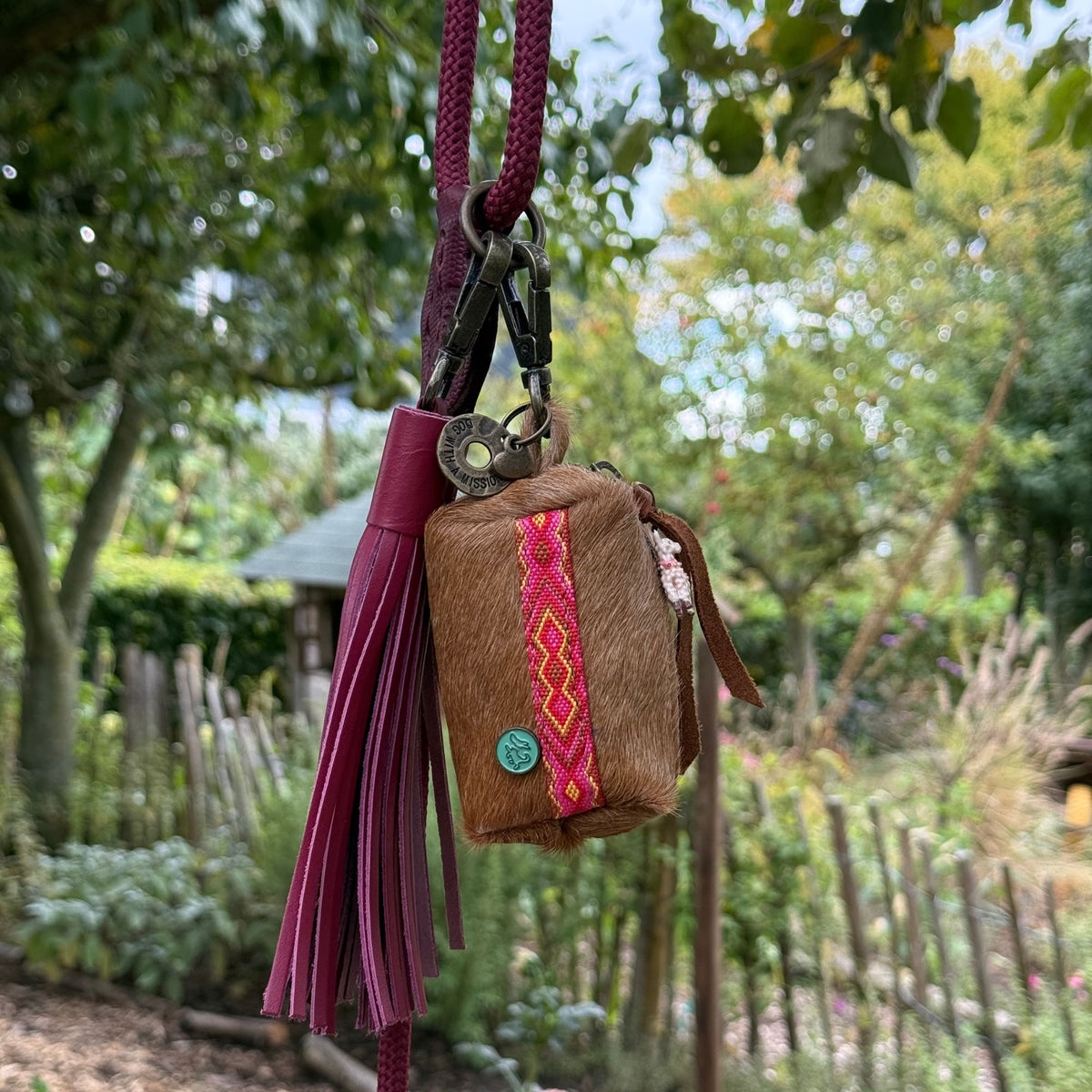 Yuki poepzakjeshouder en bordeauxrode charm hangen aan een bordeauxrode hondenriem in een tuin